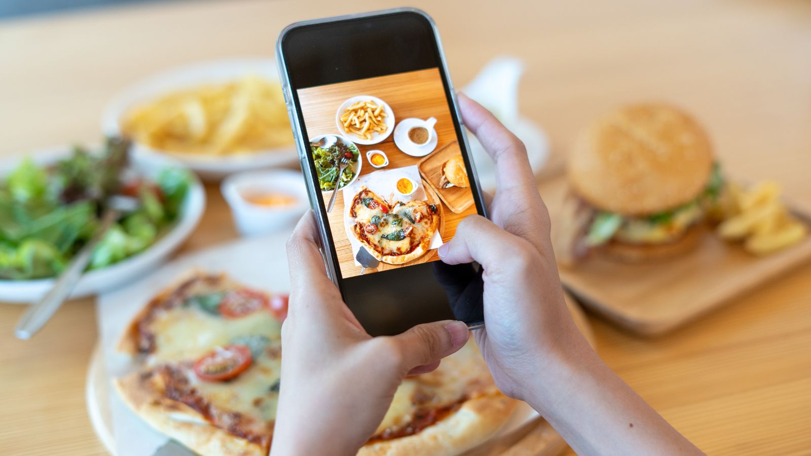 Manos sosteniendo un teléfono que fotografía una mesa con pizza, ensalada, papas fritas y una hamburguesa