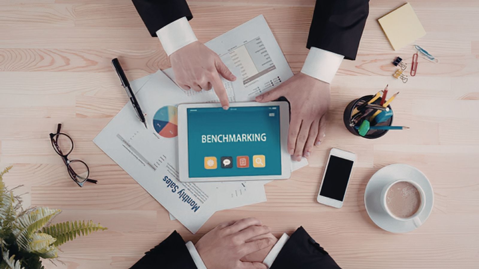 Vista cenital de un escritorio con dos personas revisando reportes impresos y una tablet que muestra la palabra benchmarking