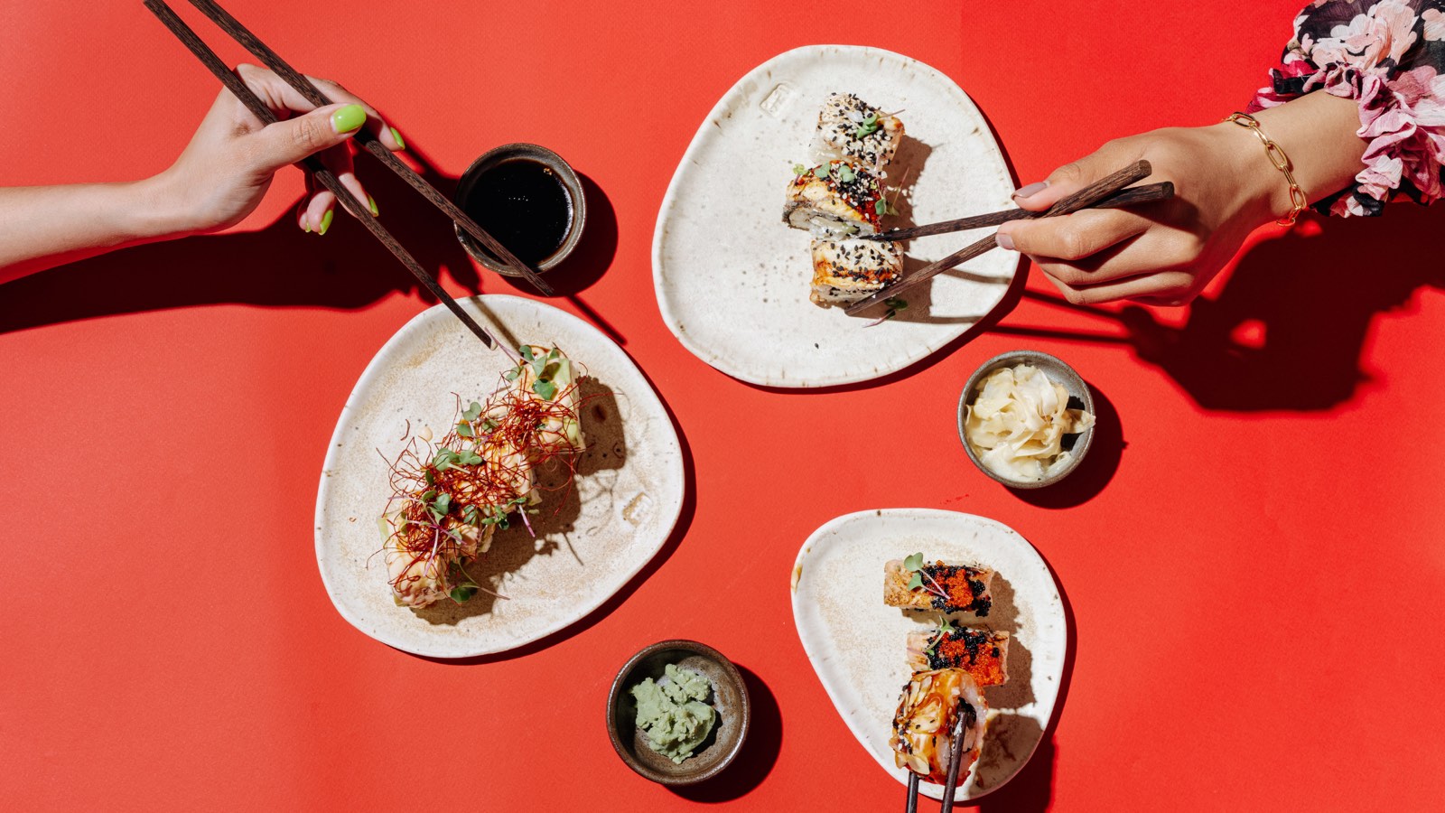 Dos personas comparten rolls de sushi con palillos, junto a salsa de soya, wasabi y jengibre