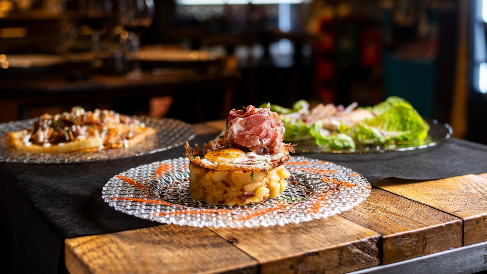 Plato de restaurante montado con papas, huevo frito y jamón sobre mesa de madera