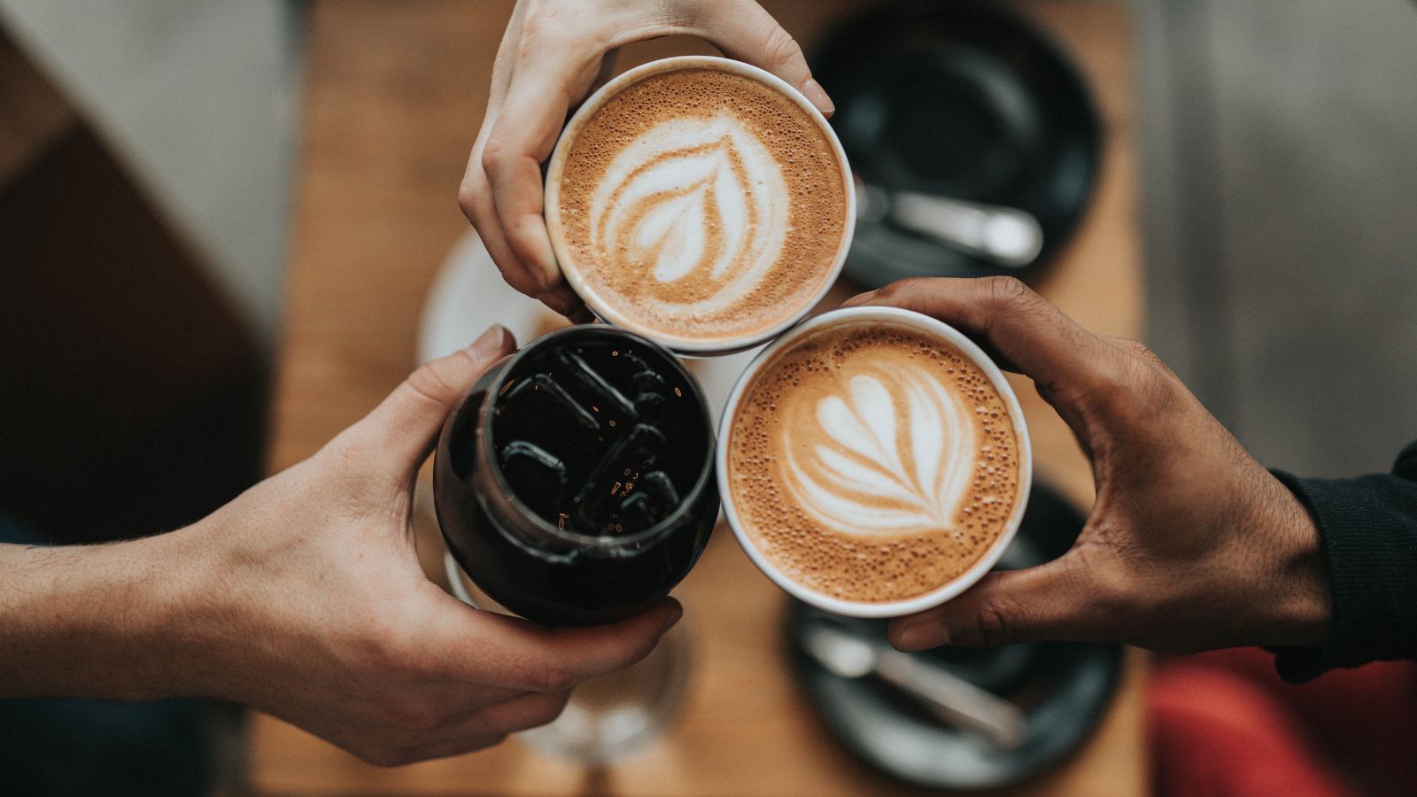Día del Café, tres manos brindando con dos capuchinos con arte latte y un café frío