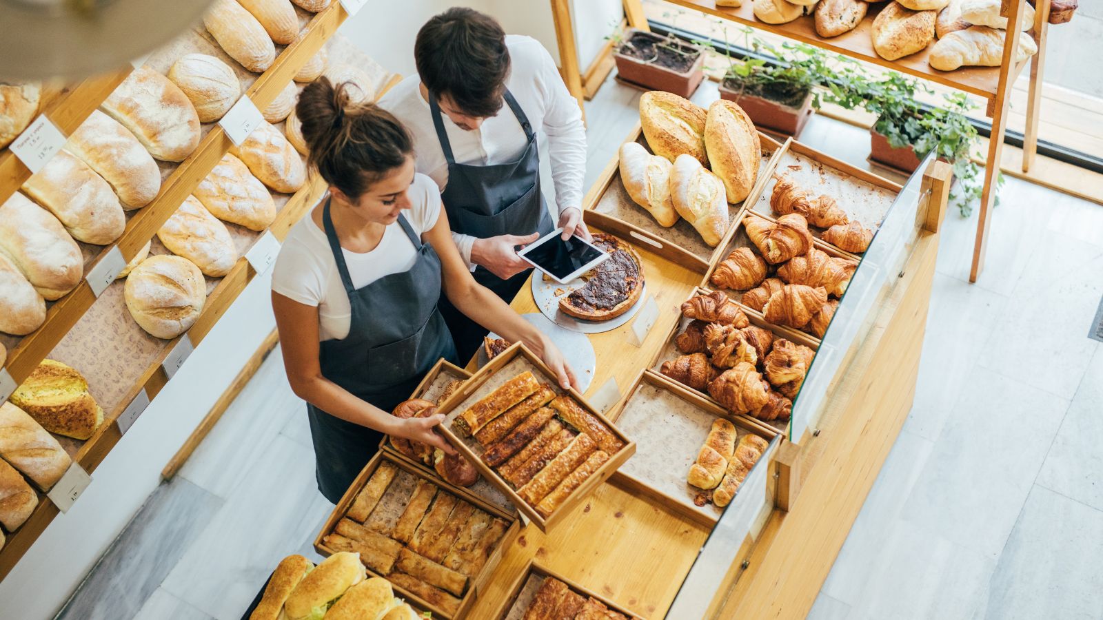 Vista cenital de una panadería con dos personas de delantal ordenando bandejas entre estantes llenos de pan y croissants